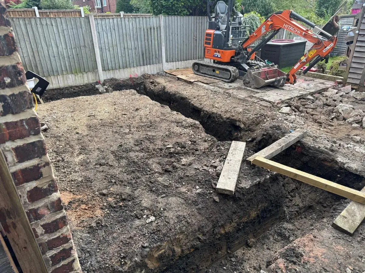 Excavator digging a service trench alongside a stone boundary wall